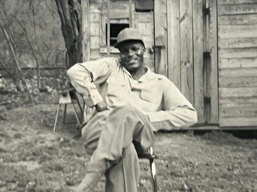 Black-and-white photograph of a man seated in a chair outdoors, smiling at the camera, resting one arm on the chair back, in front of the weathered clapboard wall of a small shed.